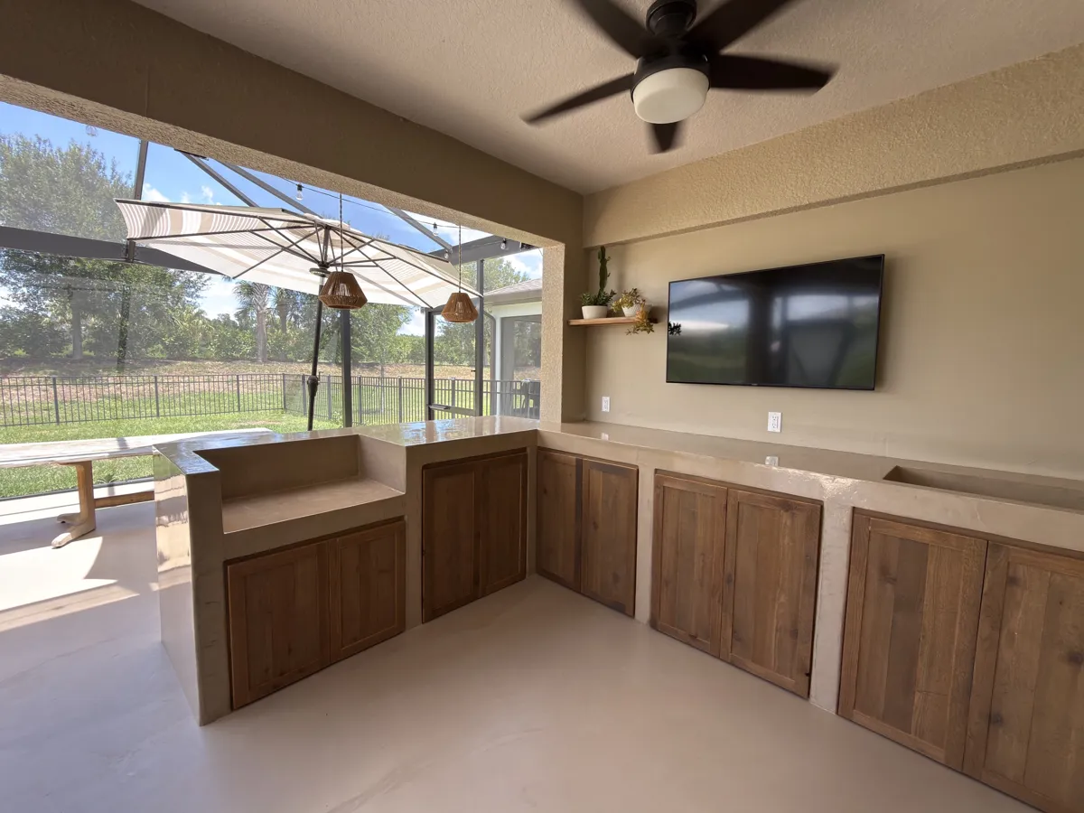 After: the outdoor kitchen finished in smooth sand-colored microcement with wood cabinet doors and a mounted TV