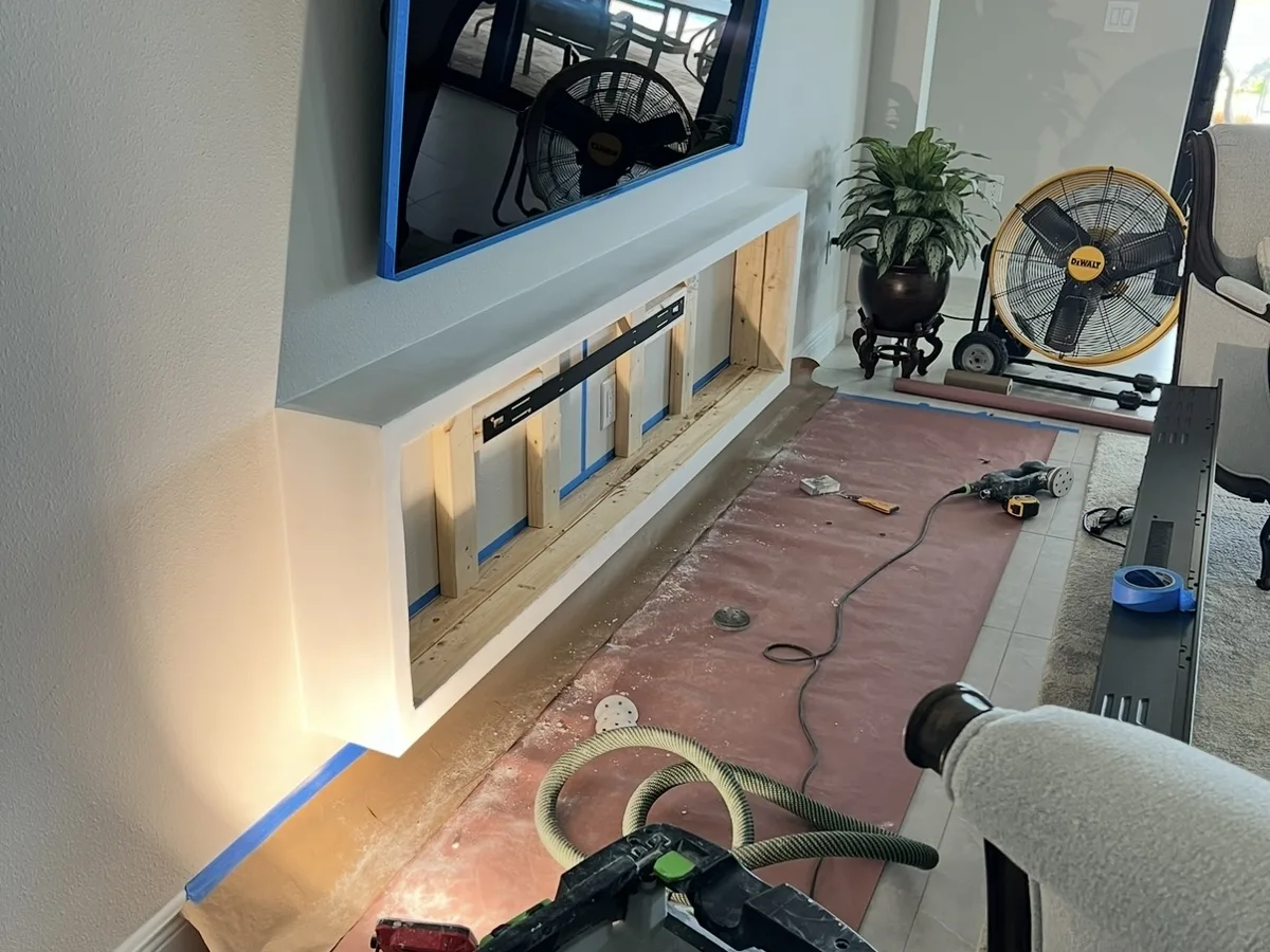 During: floating fireplace box framed in wood under a wall-mounted TV, area masked and a dust-extractor vacuum on site