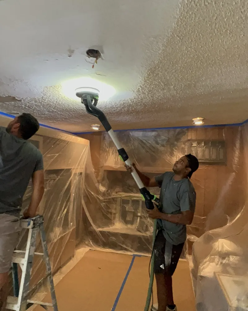 Crew removing popcorn ceiling texture with a dust-extracting sander, room fully sealed in plastic