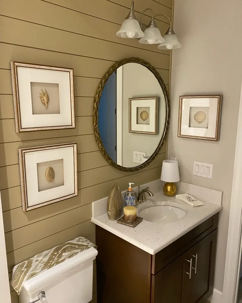 After: the half bath shiplap wall painted a warm khaki with a round brass mirror and framed shell art