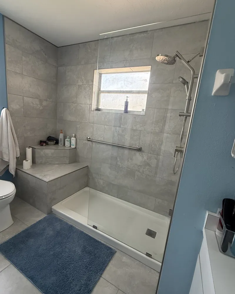 After: walk-in shower with large gray tile, tiled corner bench, glass panel, rain head with handheld and grab bar