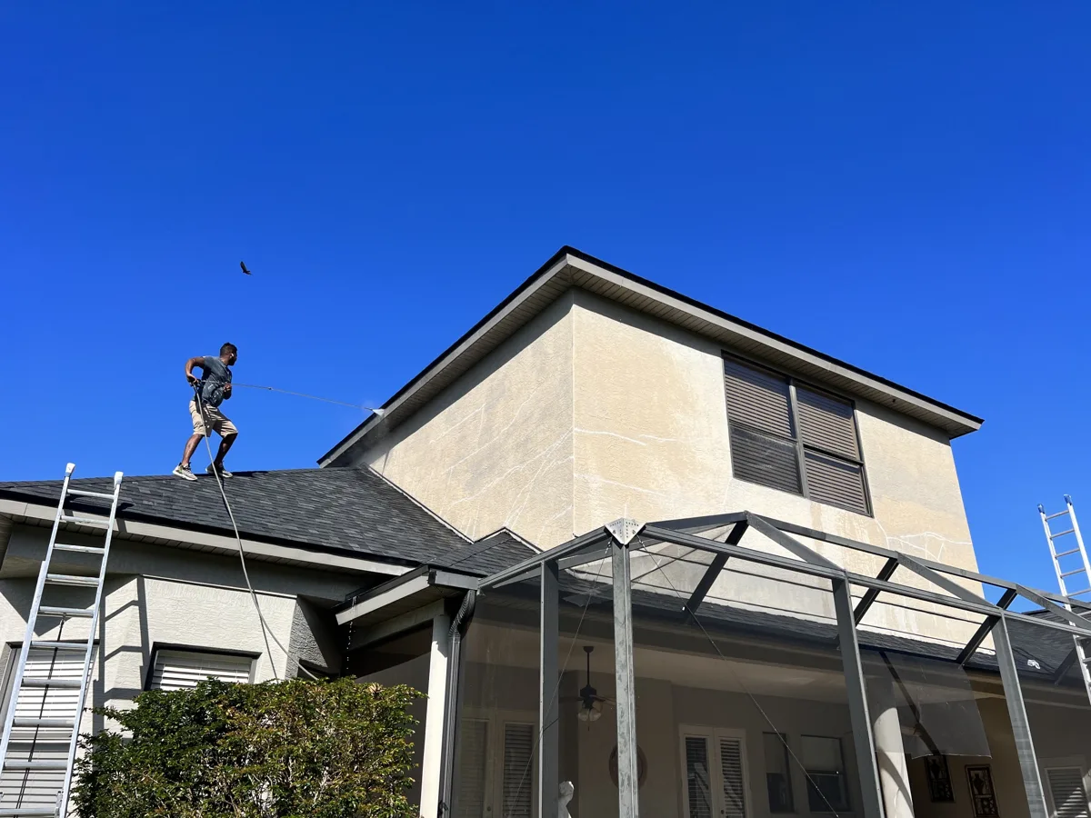 Before: crew member on the roof pressure washing a two-story stucco wall with visible patched cracks