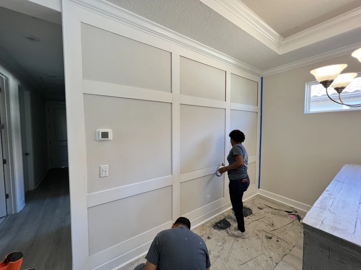 After: floor-to-ceiling board-and-batten grid installed on the same wall while the crew finishes caulking and taping