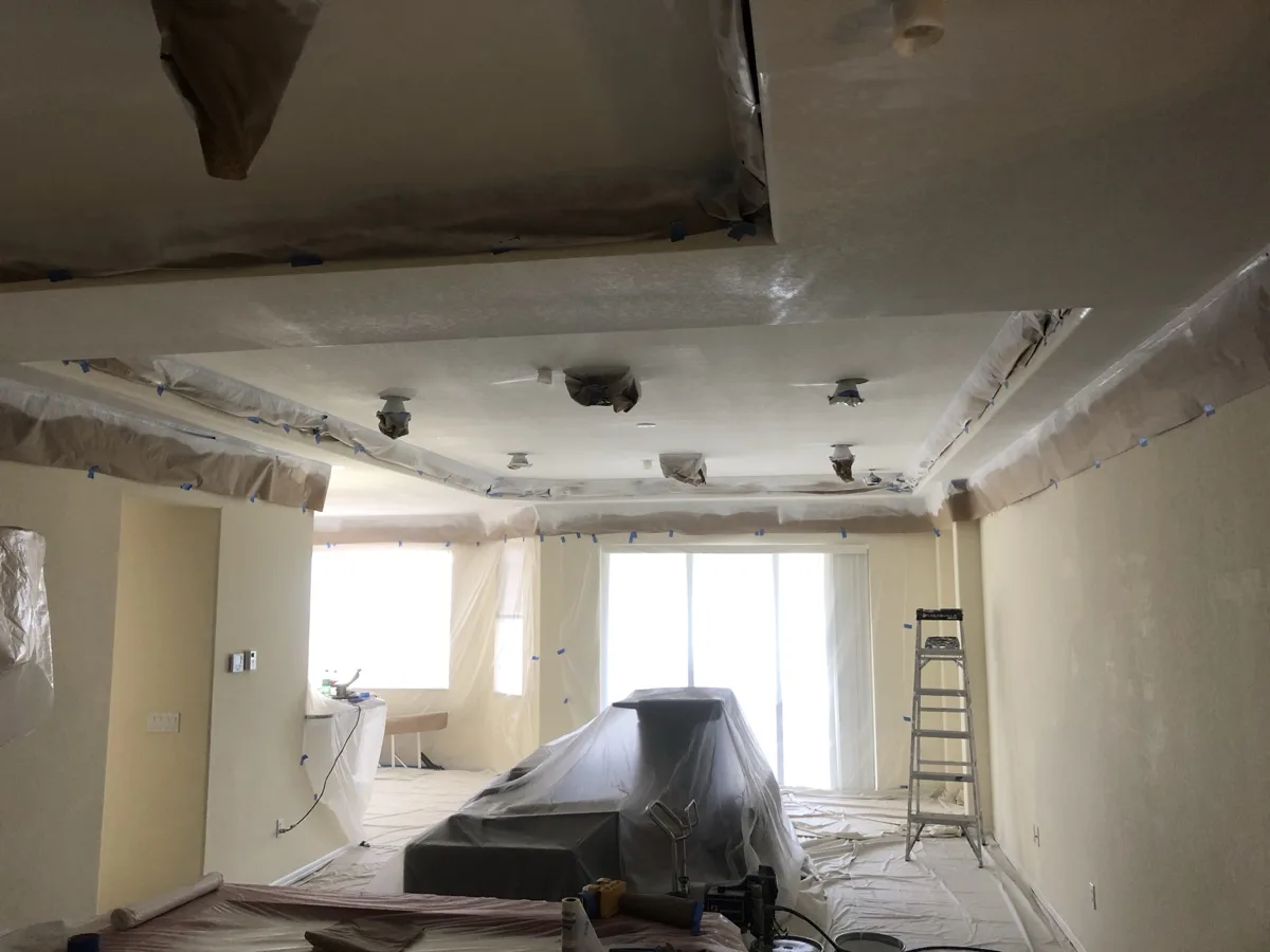 During: open living area fully masked with plastic and paper, ceiling being sprayed